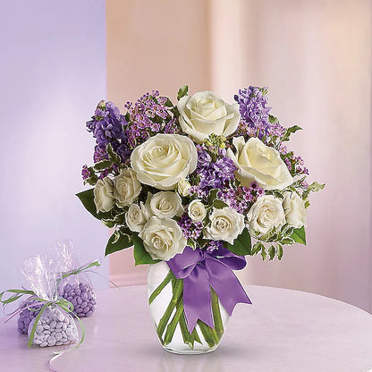 Elegant floral arrangement of white roses and purple blooms in a transparent vase with a purple bow