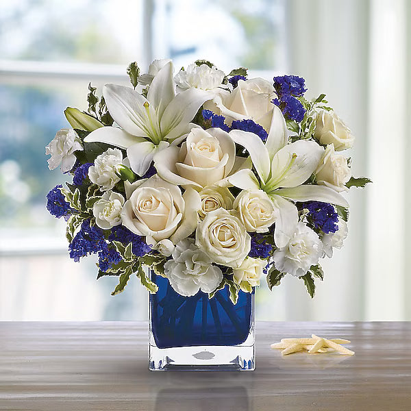 Bouquet of white and blue flowers in a clear vase on a wooden surface with a blurred window background.